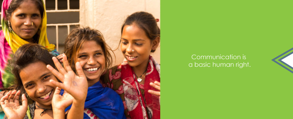 Three young girls and an older woman in colorful clothing smile and wave at the camera. Text says, "Communication is a basic human right."