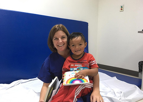 Speech pathologist Kate sits with a young client, who is holding a book, in her lap. Both are smiling.