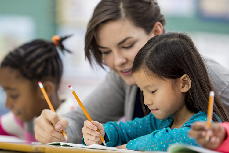 A multi-ethnic group of elementary school children are indoors in a classroom. They are wearing casual clothing. The students are sitting at their desks and writing with pencils. Their teacher is helping an Asian woman.